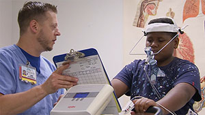 A hospital tech (left) holds a clipboard as a boy in testing equipment on a bike machine.
