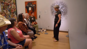 A woman (right) stands in front of a group of people and discusses the art behind her.