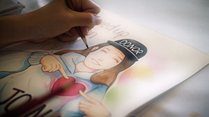 Close-up of a person's left hand drawing a smiling girl in a baseball cap