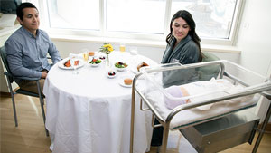 A man and a woman sit at a table with food on it while looking at a bassinet with a baby in it.