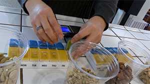 A pair of hands places nuts into a pill-a-day container on a tile counter; in front is a plastic cup with nuts in it.