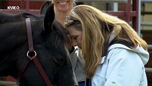 Image of a horse (left) and a woman (right) pressing their foreheads together while another person smiles in the background