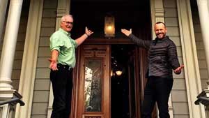 Two men stand in front of the McHenry Mansion museum entrance, hands outstretched to show off the door.