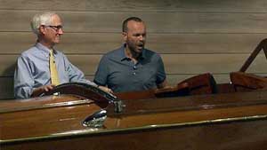 Two men stand behind a vintage wooden boat.
