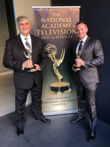 Two men in suits (Toby Momtaz, left; Tyler Bastine, right) each hold an Emmy statue on either side of a banner for the National Academy of Television Arts & Sciences