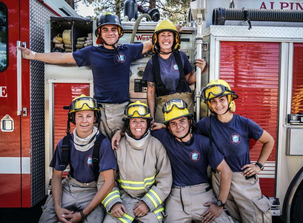 Six college students in firefighting gear pose in front of a firetruck.