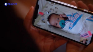 A woman holds a phone displaying a picture of a baby wrapped in a blanket with a pacifier in a hospital crib.