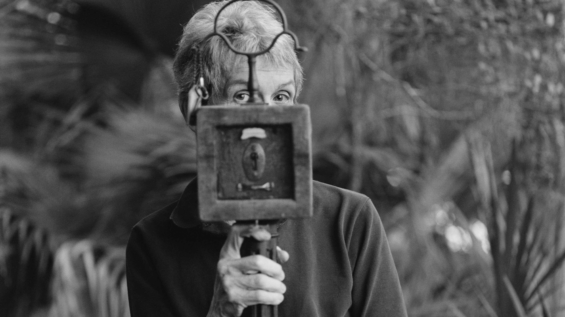 A black and white photograph of a woman posing behind a mixed-media sculpture of a framed panel with a lock and metal fixtures, her eyes peering over the top of the frame.