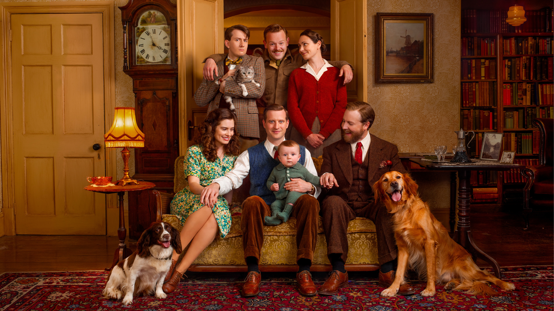 The residents of Skeldale House posing for a family photo in front of the grandfather clock and a bookshelf. Top row: Carmody, Tristan, and Mrs. Hall. Tristan has his arms around the other two, and Carmody is holding Oscar the cat. Middle row: Helen, James, baby Jimmy, and Siegfried. Baby Jimmy is sitting on James's lap. Bottom row: Dash, a little spaniel, and Jess, a big golden retriever. 